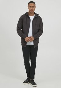 Dark gray zip-up hoodie with drawstrings, paired with a white t-shirt and black pants. Black sneakers complete the outfit. Smooth texture.
