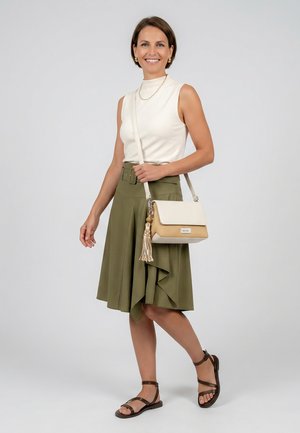 Hymyilevä nainen, jolla on päällään valkoinen hihaton toppi, oliivinvihreä polven mittainen hame, ruskeat sandaalit ja beige olkalaukku tupsu koristeella.