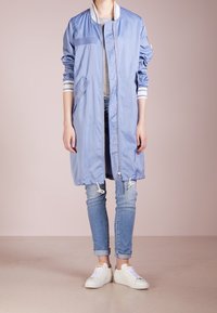 Chaqueta bomber larga color azul claro con puños de rayas blancas, cierre de cremallera, dos bolsillos laterales y bajo ajustable. Llevada sobre una camiseta gris y jeans.