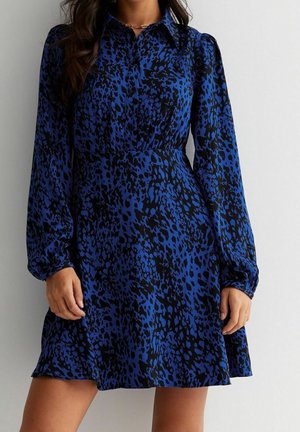 Vrouw die een blauwe en zwarte jurk met lange mouwen draagt, met dierenprint, een kraag en een getailleerde taille.