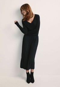 Czarny sweter z długimi rękawami i dekoltem w kształcie litery V, połączony z plisowaną czarną midi spódnicą oraz czarnymi botkami za kostkę. Gładka tekstura na całej powierzchni.