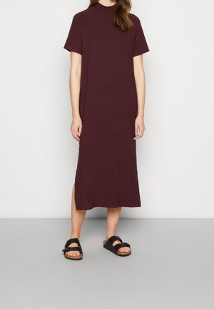 Burgunderrotes Midi-Kleid mit kurzen Ärmeln und hohem Kragen aus weichem Stoff; verfügt über seitliche Schlitze, kombiniert mit schwarzen Sandalen mit zwei Riemen.