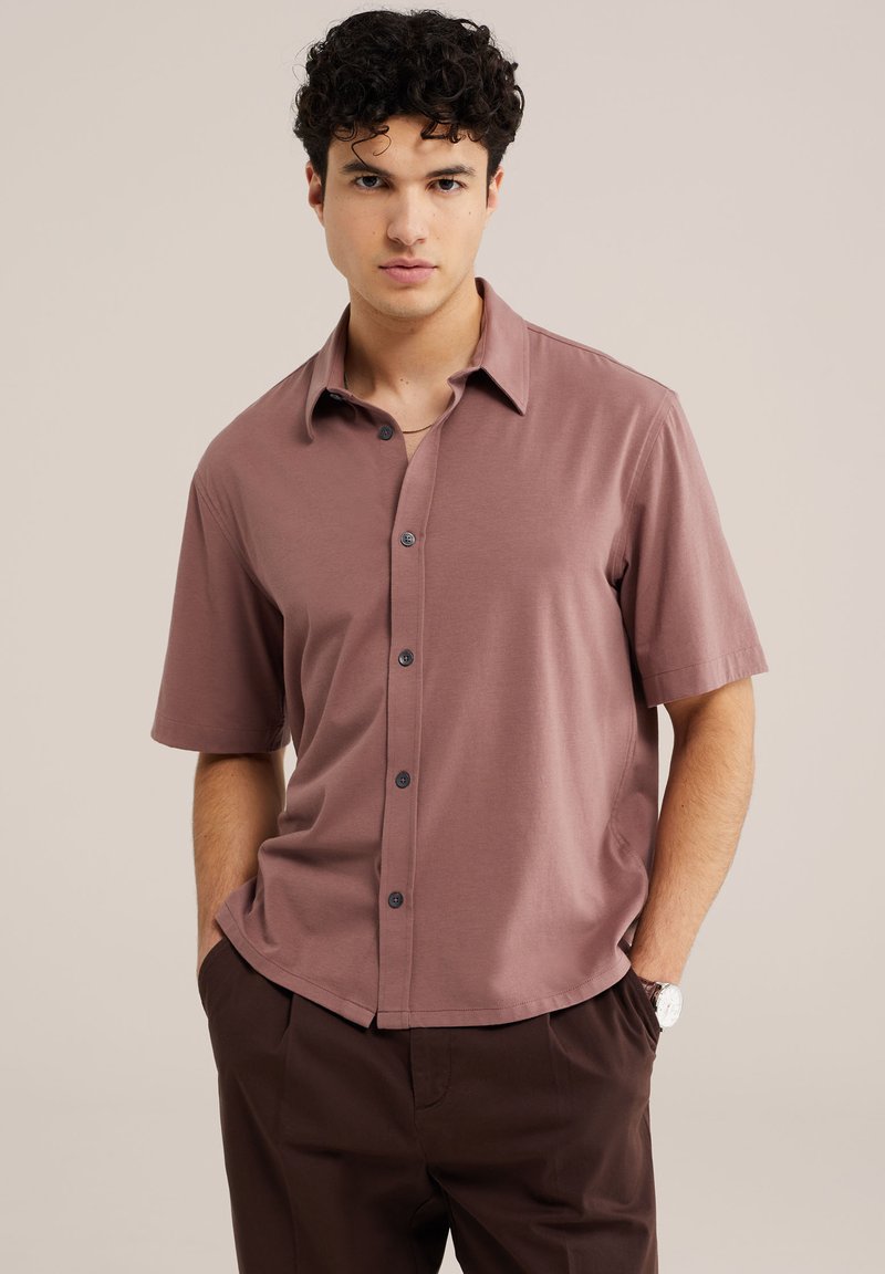 Jeune homme aux cheveux bouclés portant une chemise mauve à manches courtes boutonnée et un pantalon marron foncé, mains dans les poches, fond neutre.
