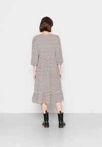 Vestido de manga longa com uma cintura franzida, apresentando um padrão geométrico repetido em roxo e verde sobre um fundo claro. Usado com botas pretas.