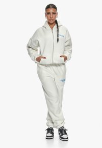 Hvit fleece hettejakke med frontglidelås og lommer, kombinert med matchende fleece joggebukser med blå broderte tekst, iført svarte joggesko.