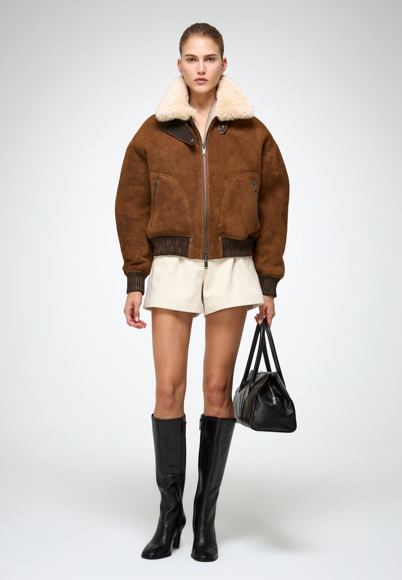 Bomber jacket in suede marrone con colletto in pelliccia, polsini a coste, due tasche frontali, abbinato a pantaloni corti beige e stivali neri in pelle alti fino al ginocchio.