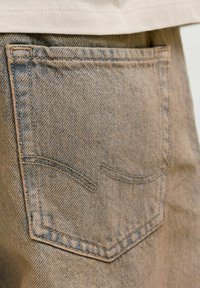 Zadní kapsa světle hnědých džín s dvojitým prošitím zakřivených linií a viditelnou texturou látky pod bílou košilí.