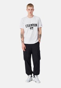 Witte katoenen t-shirt met "CHAMPION" in het zwart gedrukt, gecombineerd met zwarte cargobroeken met zijzakken en witte sneakers.
