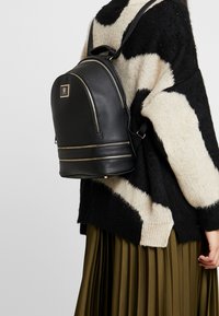 Czarna skórzana plecak z zaokrąglonym dnem, złotymi akcentami metalowymi i zamkami błyskawicznymi. Noszony na teksturowanym, paskowanym swetrze.