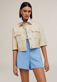 Giacca cropped in faux leather crema con due tasche sul petto e bottoni anteriori, abbinata a pantaloni shorts blu chiaro. Collare della camicia a righe visibile.