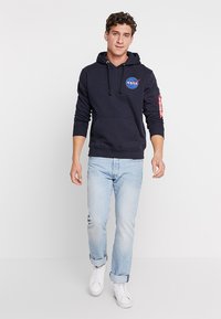 Sweatshirt cu glugă de culoare bleumarin cu logo NASA, buzunar tip kangaroo în față și buzunar cu fermoar pe mânecă cu accent roșu. Se asortează cu blugi deschiși și adidași albi.