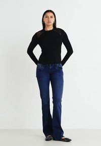 Črn dolg rokav z čipkastimi detajli, v kombinaciji z modrimi širokimi jeansami. Model nosi črne balerinke s studenci. Preprosta, čista ozadje.
