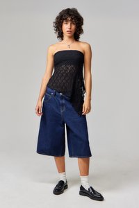 Čierny krajkový top bez ramienok s textúrovaným vzorom, spárovaný s tmavomodrými širokými denims šortkami a čiernymi loafers obuvou. Model stojí pred neutrálnym pozadím.