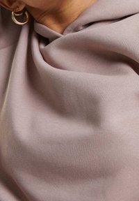 Primer plano de una persona que lleva una camisa de tela malva con pendientes de aro dorados, mostrando el área del cuello y los hombros.