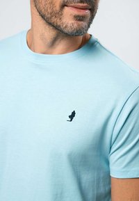 Vyras dėvi šviesiai mėlyną marškinėlį su mažyčiu juodu siuvinėjimu kairėje krūtinės pusėje – vaikščiojančio žmogaus figūra, laikanti portfelį.