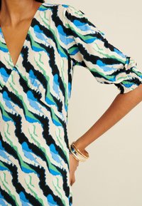 Sukienka z falistym wzorem w kolorze czarnym, niebieskim i zielonym na jasnobeżowym tle, z bufkami na rękawach i głębokim dekoltem w kształcie litery V.
