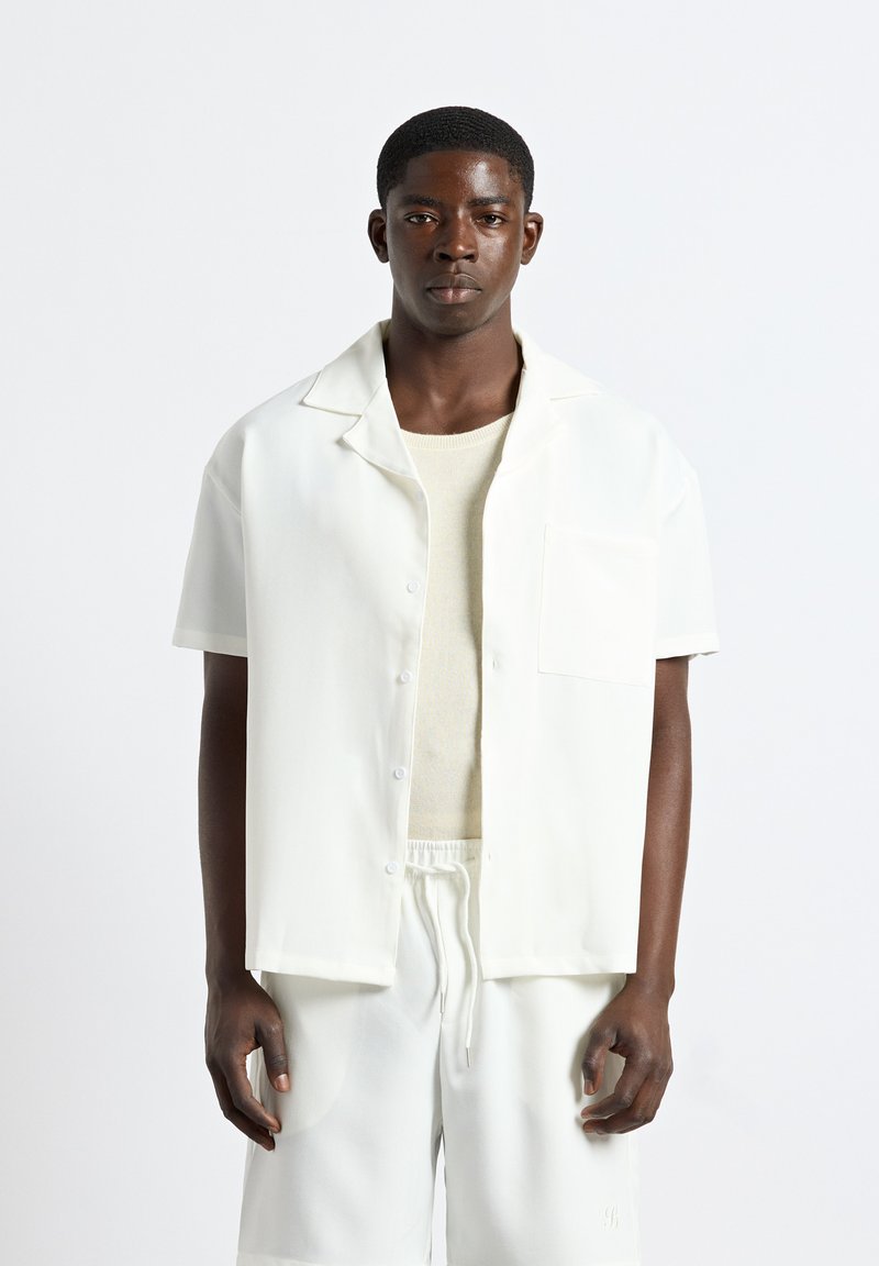 Jeune homme portant une chemise blanche unie à manches courtes boutonnée par-dessus un pull léger et un short blanc assorti avec taille à cordon.