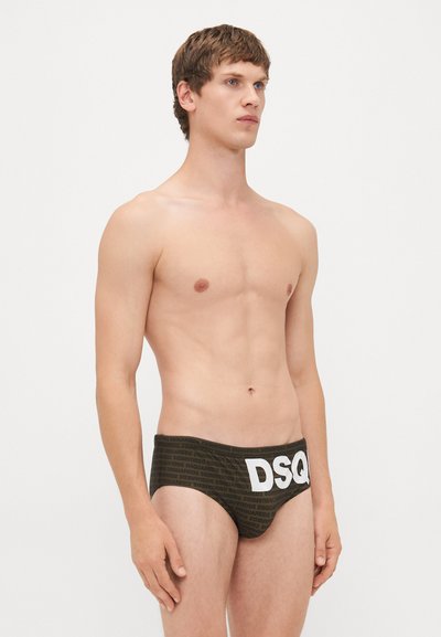 Mlad moški s kratkimi lasmi, oblečen v temne hlačke z večkratnim napisom "DSQUARED2" in velikim belim "DSQ" spredaj, stoji ob belem ozadju.