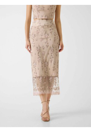 Jupe midi beige à fleurs brodées avec ourlet transparent, portée avec un haut assorti et des sandales à talons hauts beiges à brides.