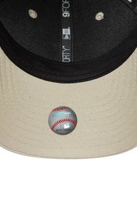 Beige Baseballcap mit schwarzem Schild, versehen mit einem silbernen Aufkleber, der wie ein Baseball aussieht, auf der Vorderseite. Strukturierte Form mit flachem Schirm.