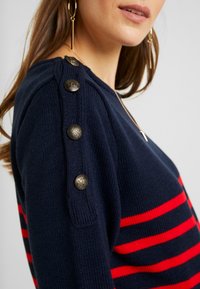 Marineblauer Strickpullover mit roten horizontalen Streifen, dekorativen Metallknöpfen an der Schulter, Rundhalsausschnitt und strukturiertem Stoff.