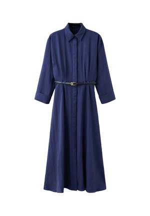 Femme debout contre un mur blanc uni, portant une longue robe bleu marine ceinturée et des escarpins noirs à bout pointu.