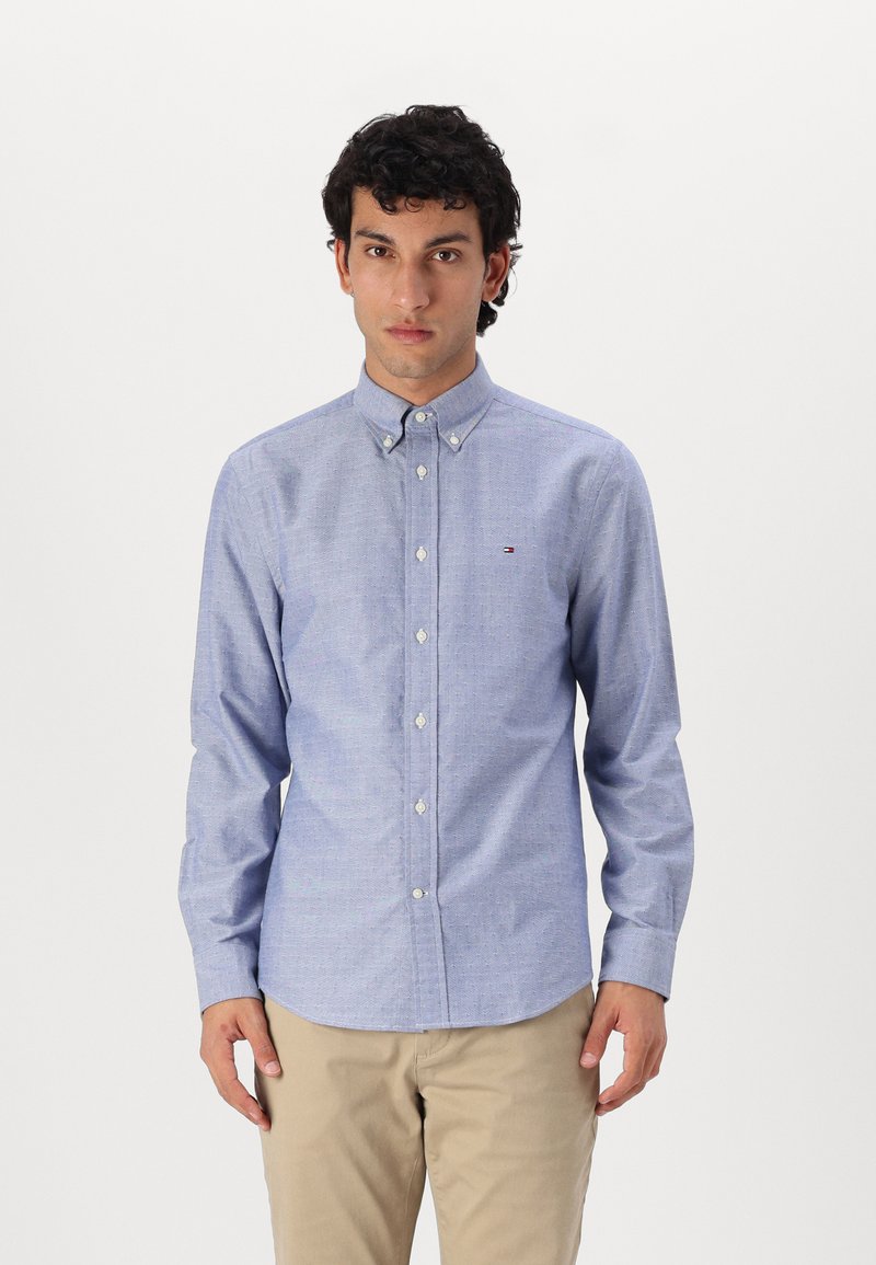 Camisa de botones azul claro con un pequeño patrón de cuadros, fabricada en algodón y con un cuello clásico. Presenta botones blancos y un logotipo en el pecho.