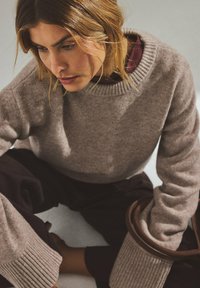 Light brown knit sweater with a crew neck and ribbed cuffs. Textured fabric sits over a maroon collared shirt, paired with dark trousers.