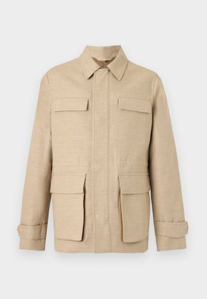 Man met kort blond haar, draagt een beige jas, een zwart shirt, donkere broek en beige-bruine loafers, staand tegen een witte achtergrond.