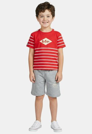 Garçon souriant portant un T-shirt Lee Cooper rayé rouge et blanc, un short gris et des baskets blanches, debout devant un fond uni.