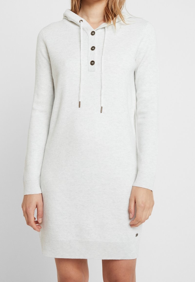 Femme portant une robe en maille gris clair à manches longues avec capuche, trois boutons foncés et des cordons au niveau de l'encolure.