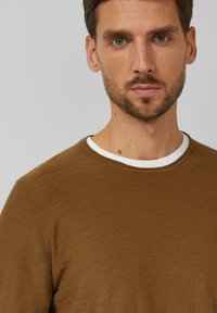 Bruine trui met ronde hals en een gestructureerde brei, voorzien van een witte binnenkraag. De stof lijkt zacht en licht van gewicht.