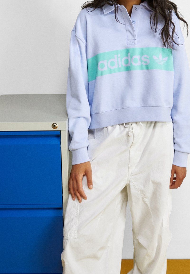 Ljust lila kort sweatshirt med en teal adidas-logotyp över framsidan, i kombination med lösa vita byxor. Mjuk tyg och avslappnad passform.
