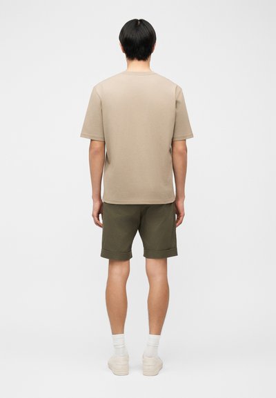 Άνδρας που στέκεται με την πλάτη, φοράει μπεζ κοντομάνικο πουκάμισο, χακί κατεβασμένα κοντά παντελόνια, λευκές κάλτσες και μπεζ αθλητικά παπούτσια σε απλό φόντο.