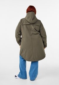 Olijfgroene lange parka met capuchon, heeft een rechte snit, middennaad en verstelbare trekkoord in de taille, gecombineerd met lichtblauwe wijde jeans.
