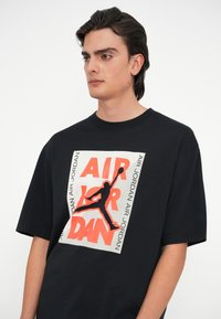 Μαύρο κοντομάνικο T-shirt με μεγάλη γραφιστική σχεδίαση που περιλαμβάνει κόκκινο και λευκό κείμενο και μια σιλουέτα καλαθοσφαιριστή σε δράση.