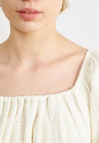 Vaalean kermaisen värinen pusero, jossa on neliömäinen pääntie. Pusero on varustettu ryppypinnalla ja pehmeällä, laskeutuvalla kankaalla. Reunoissa on yksityiskohtina röyhelöitä.