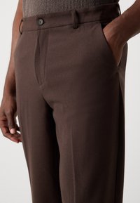 Pantalon marron en tissu lisse avec fermeture à bouton, devant plat élégant, poches latérales et coupe ajustée.
