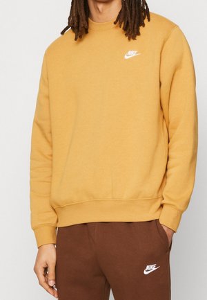 Personne portant un sweat-shirt Nike jaune moutarde et un pantalon de survêtement Nike marron, debout avec une main dans la poche sur un fond uni.