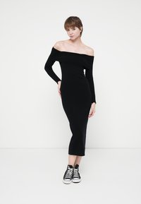 Rochie neagră, cu umăr gol, ajustată pe corp, lungime medie până la glezne, mâneci lungi, asortată cu teniși negri și albi, dintr-un textile neted.