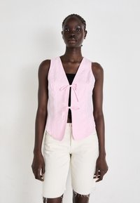 Colete de linho rosa com decote em V profundo, apresentando um fecho frontal com laço e bainha pontiaguda, combinado com calções de ganga claros e desgastados.