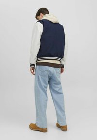 Marineblauwe en witte varsityjack met gestreepte manchetten, gelaagd over een beige hoodie, in combinatie met lichtblauwe losse jeans en tan kleurige laarzen.