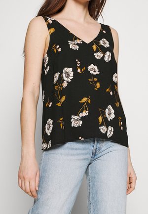 Kvinde iført ærmeløs sort top med hvidt og brunt blomsterprint og lyseblå jeans, stående mod en ensfarvet baggrund.