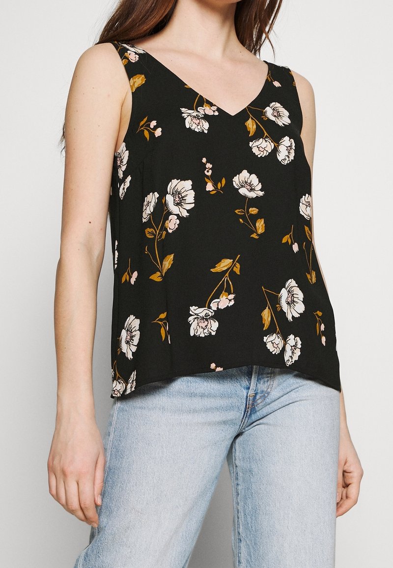 Femme portant un haut sans manches noir avec un imprimé floral blanc et marron et un jean bleu clair, debout devant un fond uni.