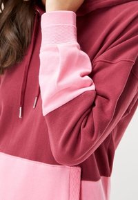 Maroon and pink color-blocked hoodie with a textured finish. Features adjustable drawstrings, ribbed cuffs, and a loose fit design.