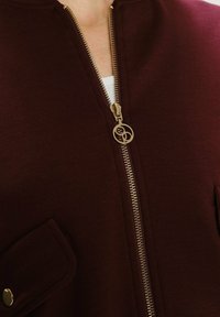 Bordeaux ritsjack met een gouden rits en een ronde trekker, voorzien van patchzakken en een zachte, gestructureerde stof.