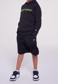 Černá mikina s kapucí se zeleným logem, černé síťované kraťasy a černo-bílé sportovní boty. Má přední kapsu a detaily s logem.