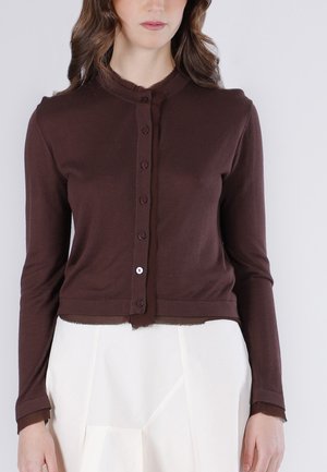 Žena v hnědém zapnutém kardiganu s opranými okraji a bílou vysokým pasem sukni na jednobarevném světlém pozadí.