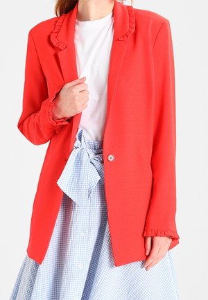 Vrouw draagt een felrode blazer met ruches over een wit overhemd en een blauwe geruite rok die in de taille met een strikje is vastgemaakt.