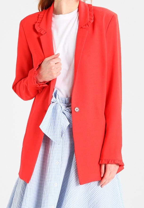 Femme portant un blazer rouge vif avec des détails à volants sur une chemise blanche et une jupe vichy bleue nouée à la taille avec un nœud.